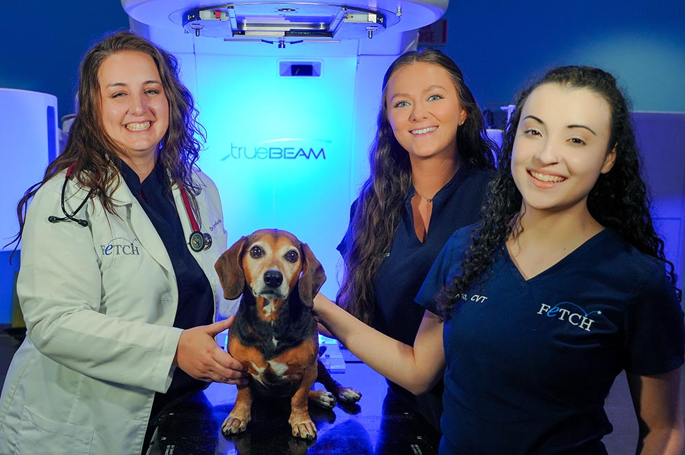 female veterinarian and two female technicians smiling and posing with small dog in front of TrueBeam machine