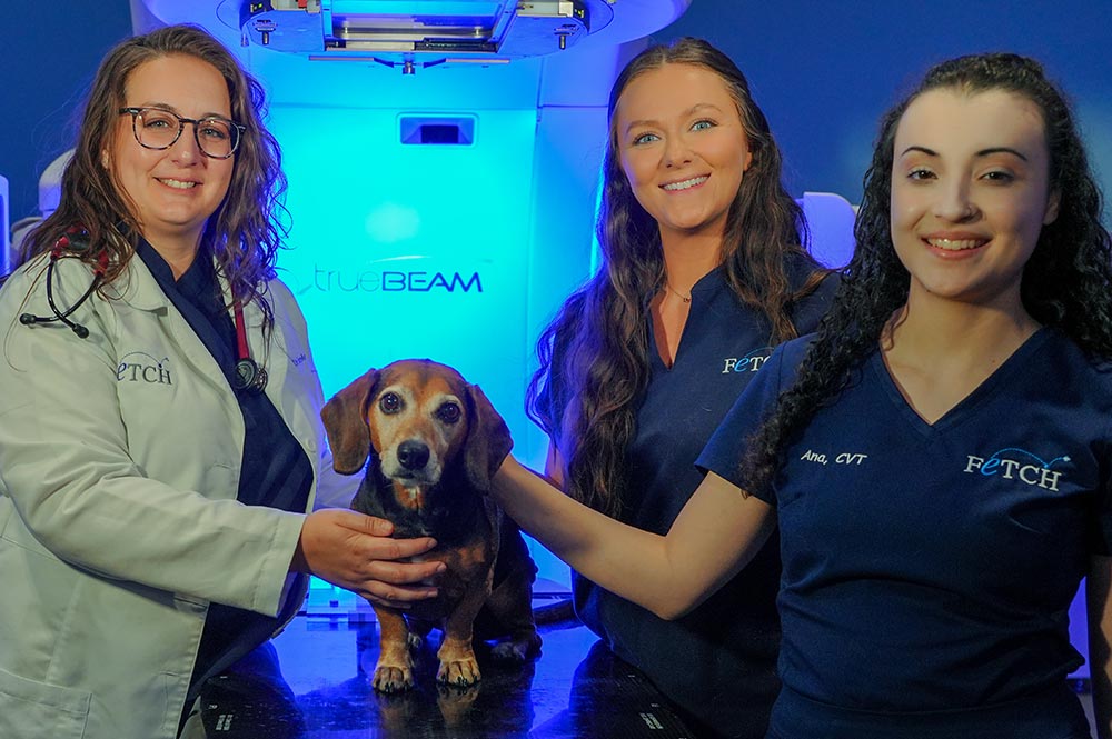 female veterinarian and two female technicians smiling and posing with small dog in front of TrueBeam machine